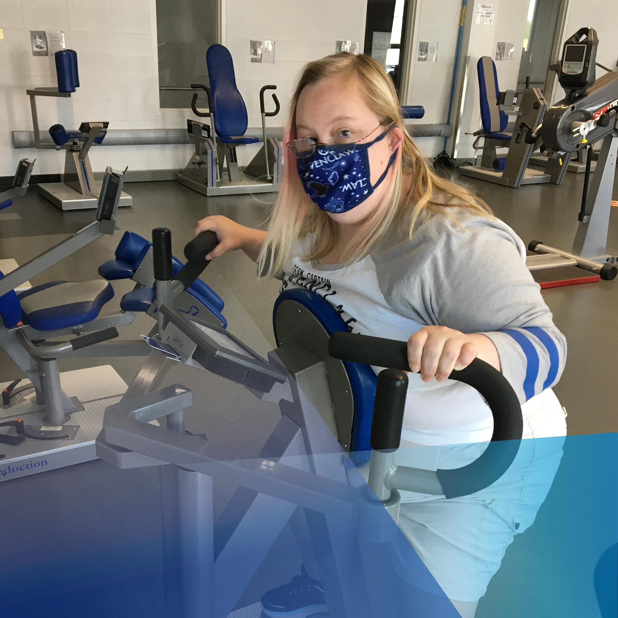 Ashley sits on a workout machine, wearing a mask, looking at the camera